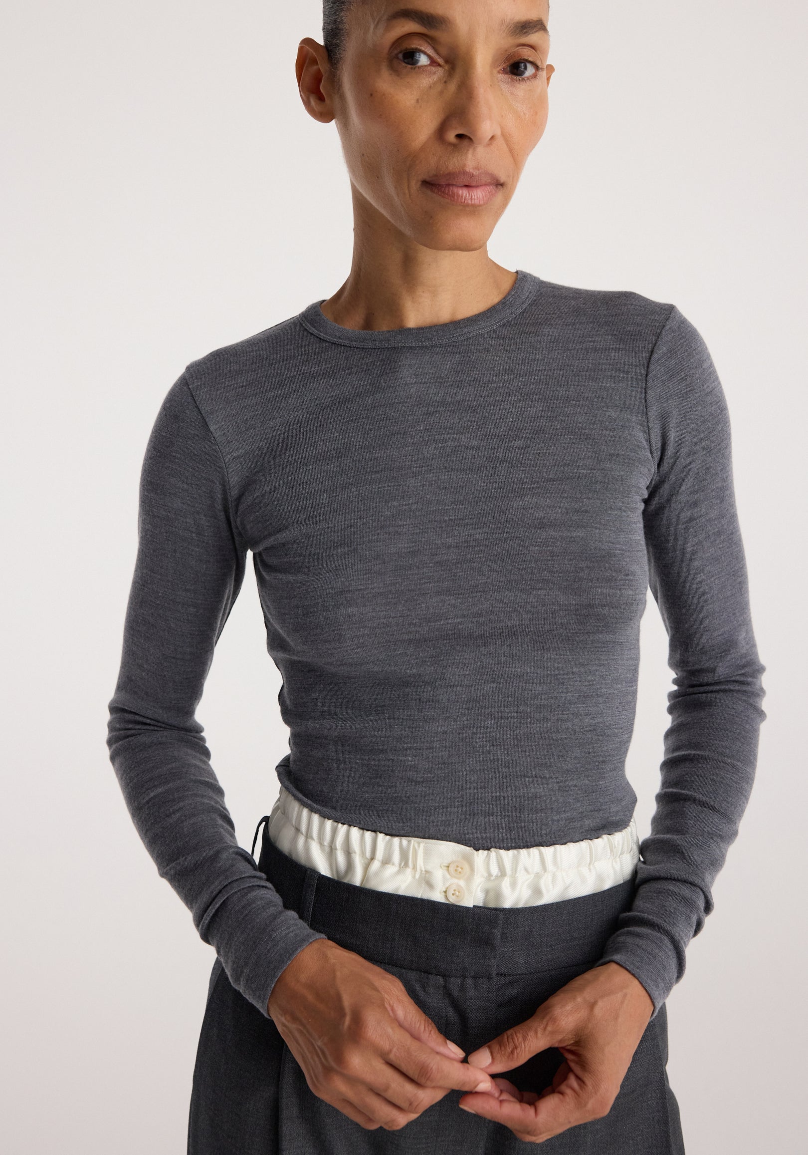 Woman wearing a fitted Fine merino long sleeve | mid grey melange shirt with long sleeves and a round neckline, paired with high-waisted pants featuring a white elastic waistband.