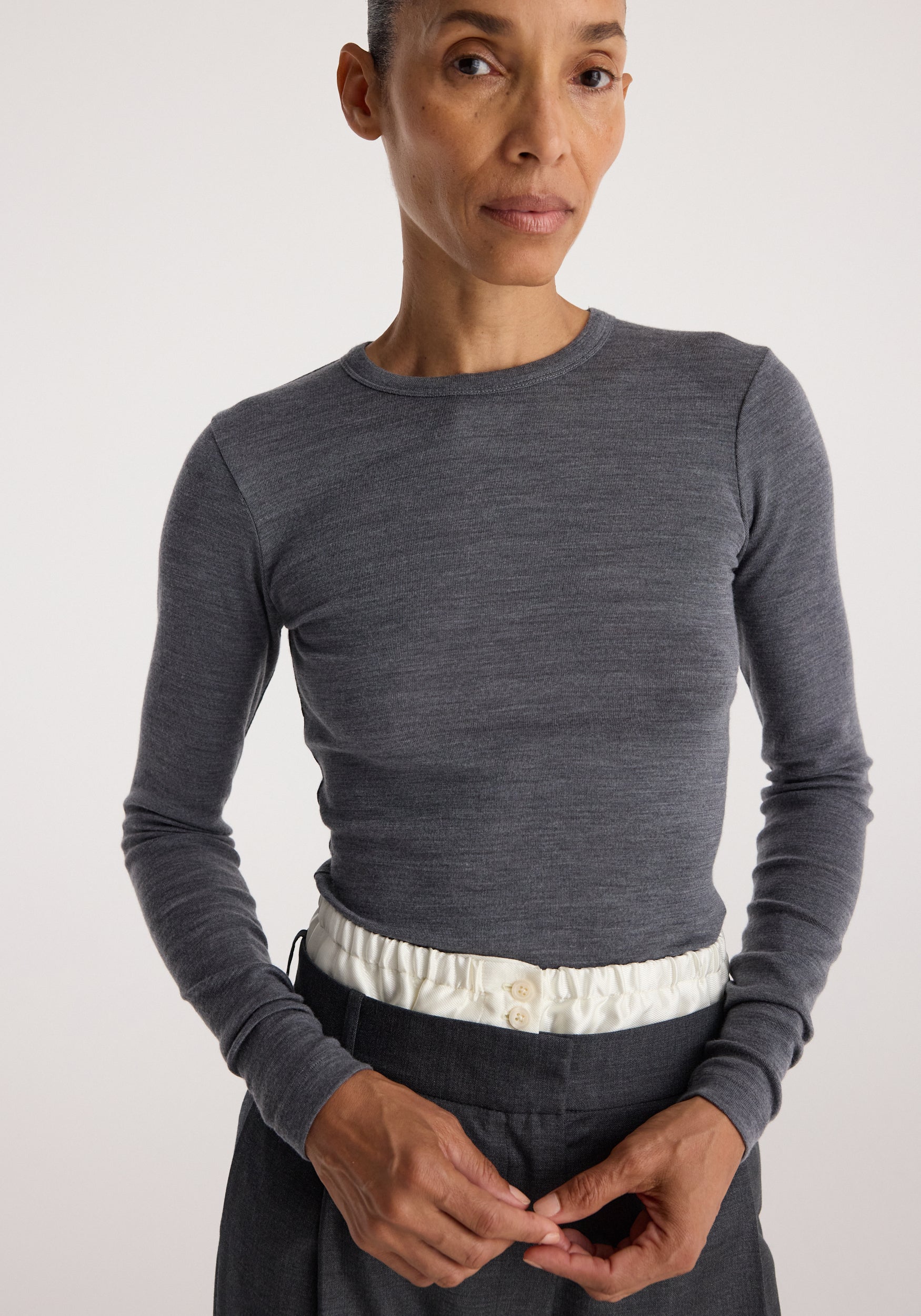 Woman wearing a fitted Fine merino long sleeve | mid grey melange shirt with long sleeves and a round neckline, paired with high-waisted pants featuring a white elastic waistband.