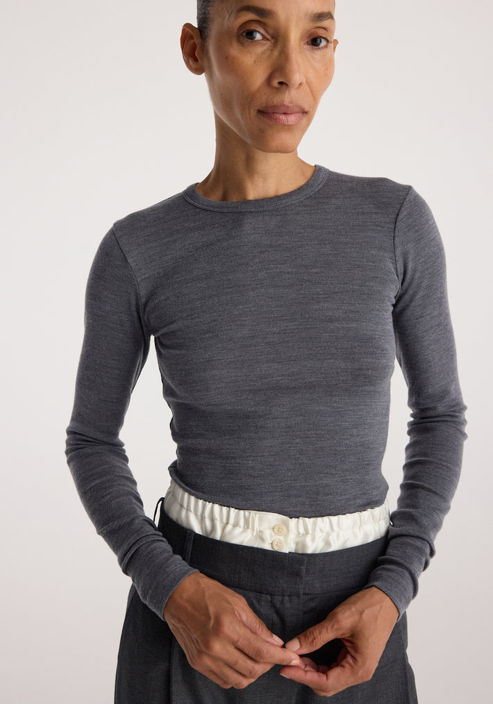 Woman wearing a fitted Fine merino long sleeve | mid grey melange top with a round neckline, paired with dark pants and a white waistband detail against a plain background.