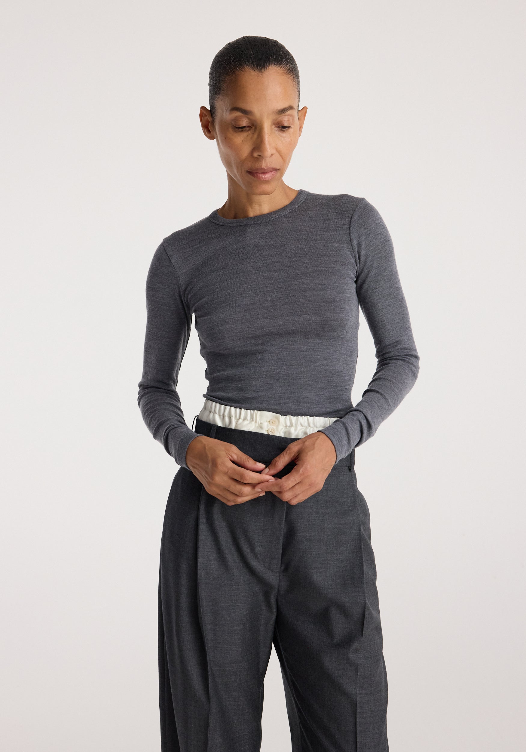 Woman wearing a mid grey melange fine merino long sleeve shirt paired with dark grey pants, standing against a plain light background with hands clasped.