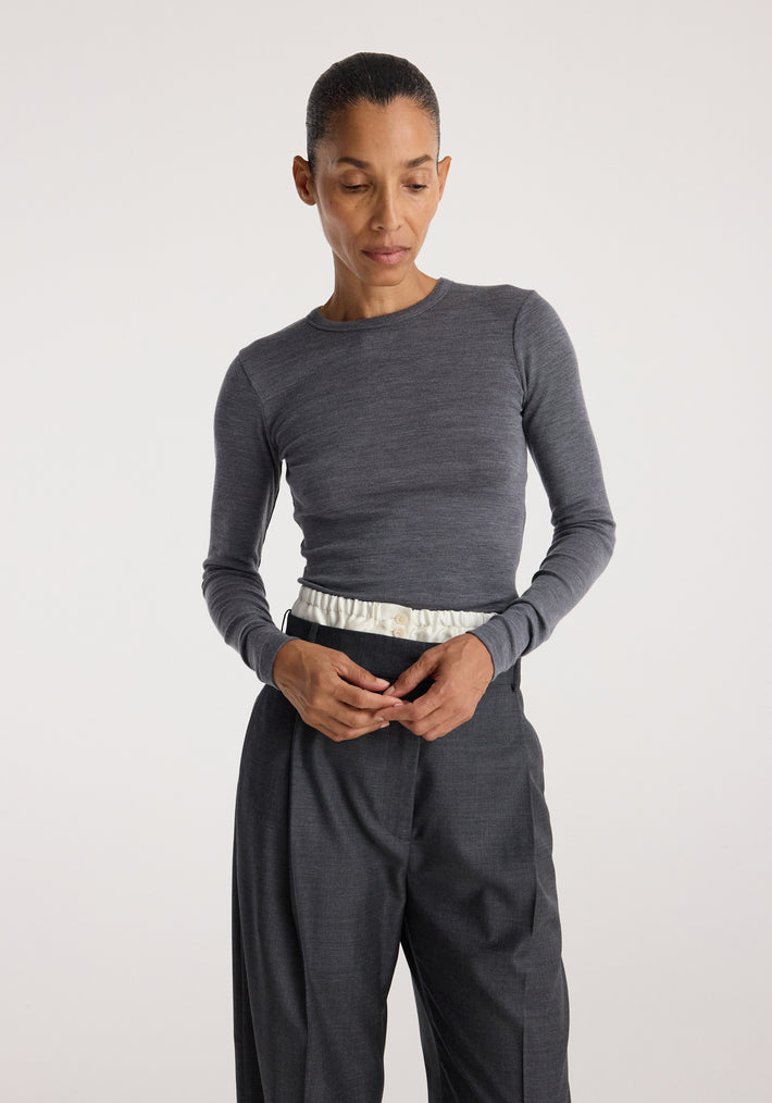 Woman wearing a mid grey melange Fine merino long sleeve top with long fitted sleeves, paired with high-waisted dark trousers against a plain background.