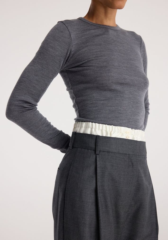 Close-up of person wearing a fitted Fine merino long sleeve | mid grey melange top paired with dark high-waisted trousers and white waistband detail against a neutral background.