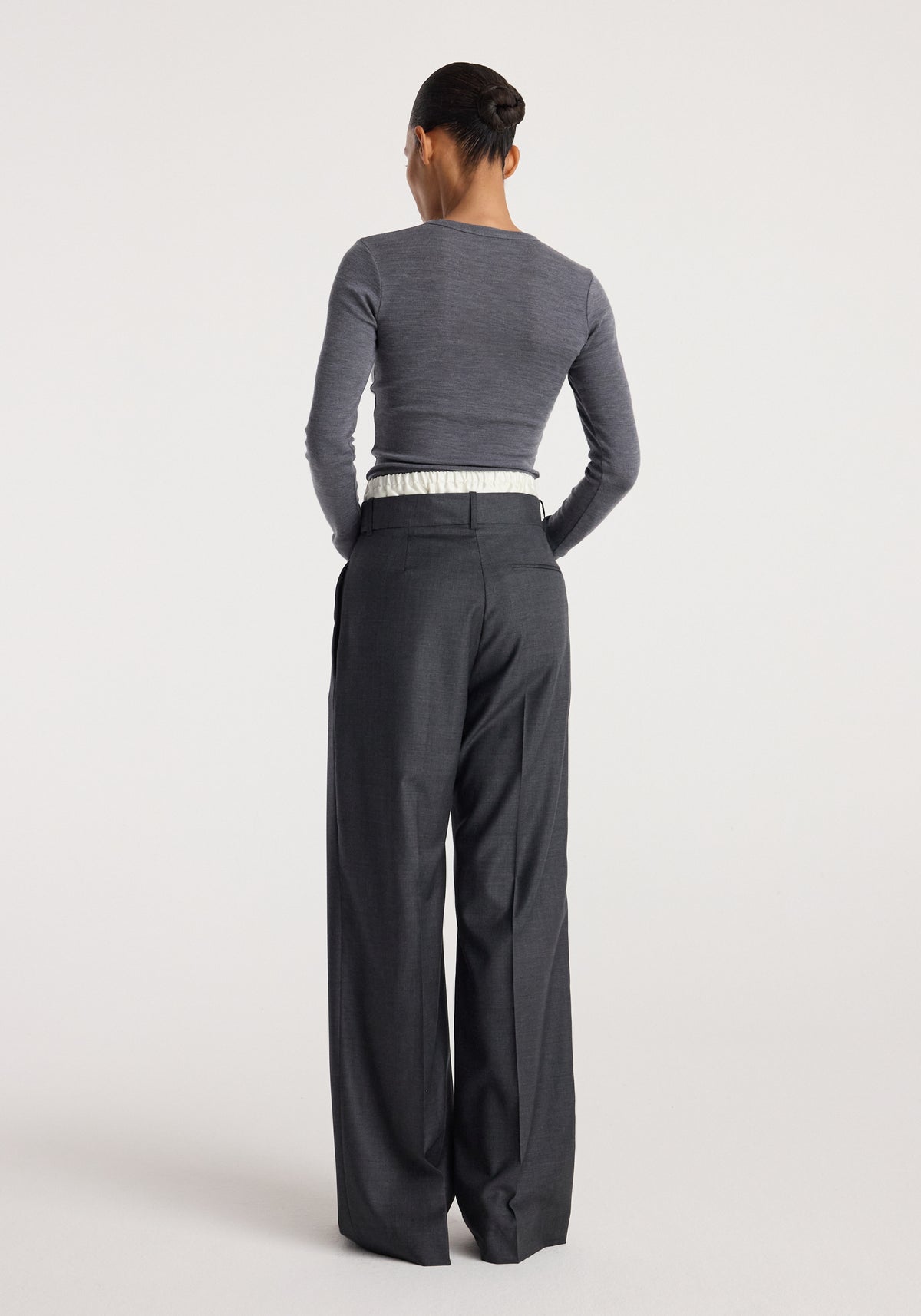 Woman seen from the back wearing a fitted mid grey melange fine merino long sleeve top paired with high-waisted dark grey wide-leg trousers against a plain background.