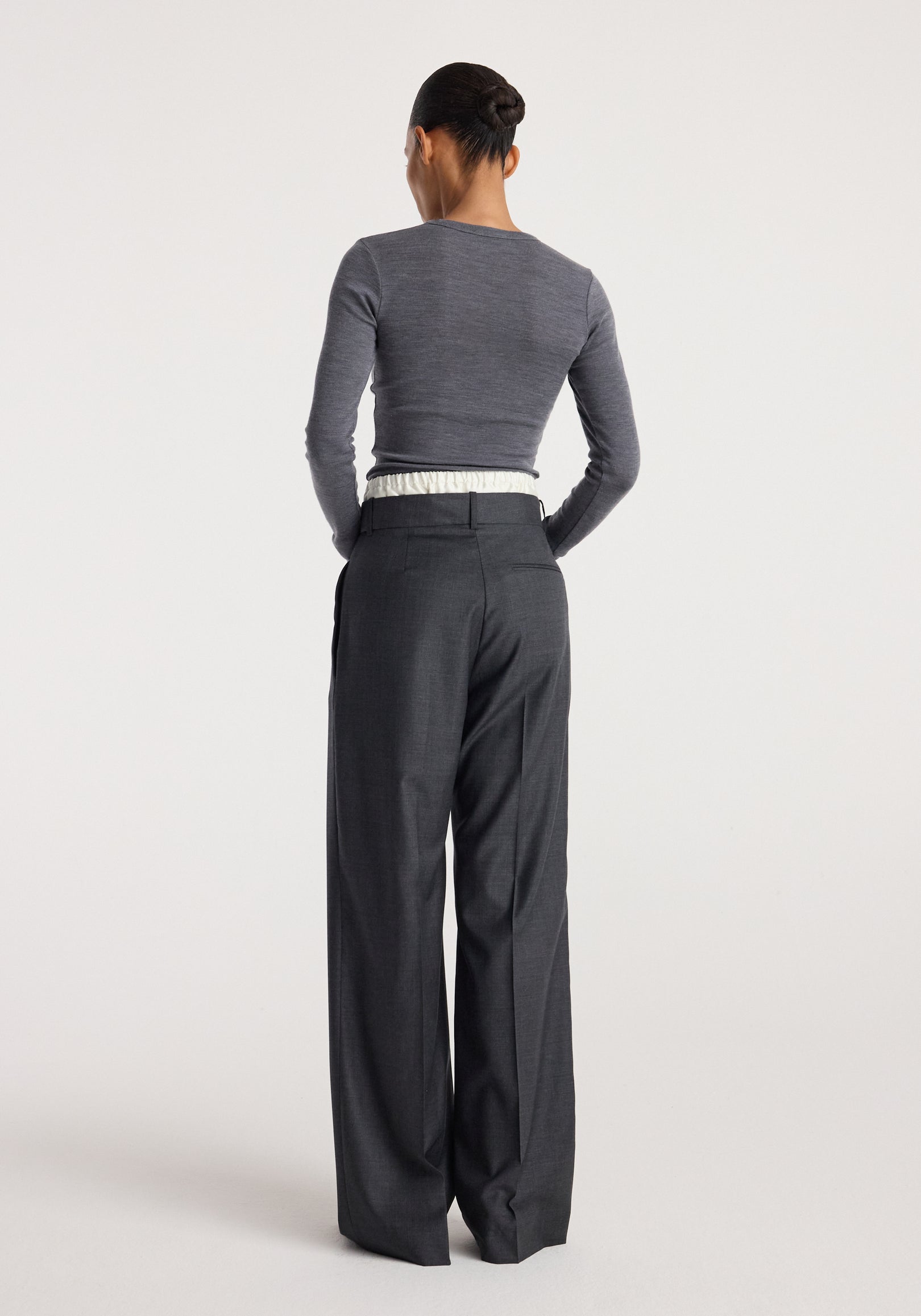 Woman seen from the back wearing a fitted mid grey melange fine merino long sleeve top paired with high-waisted dark grey wide-leg trousers against a plain background.