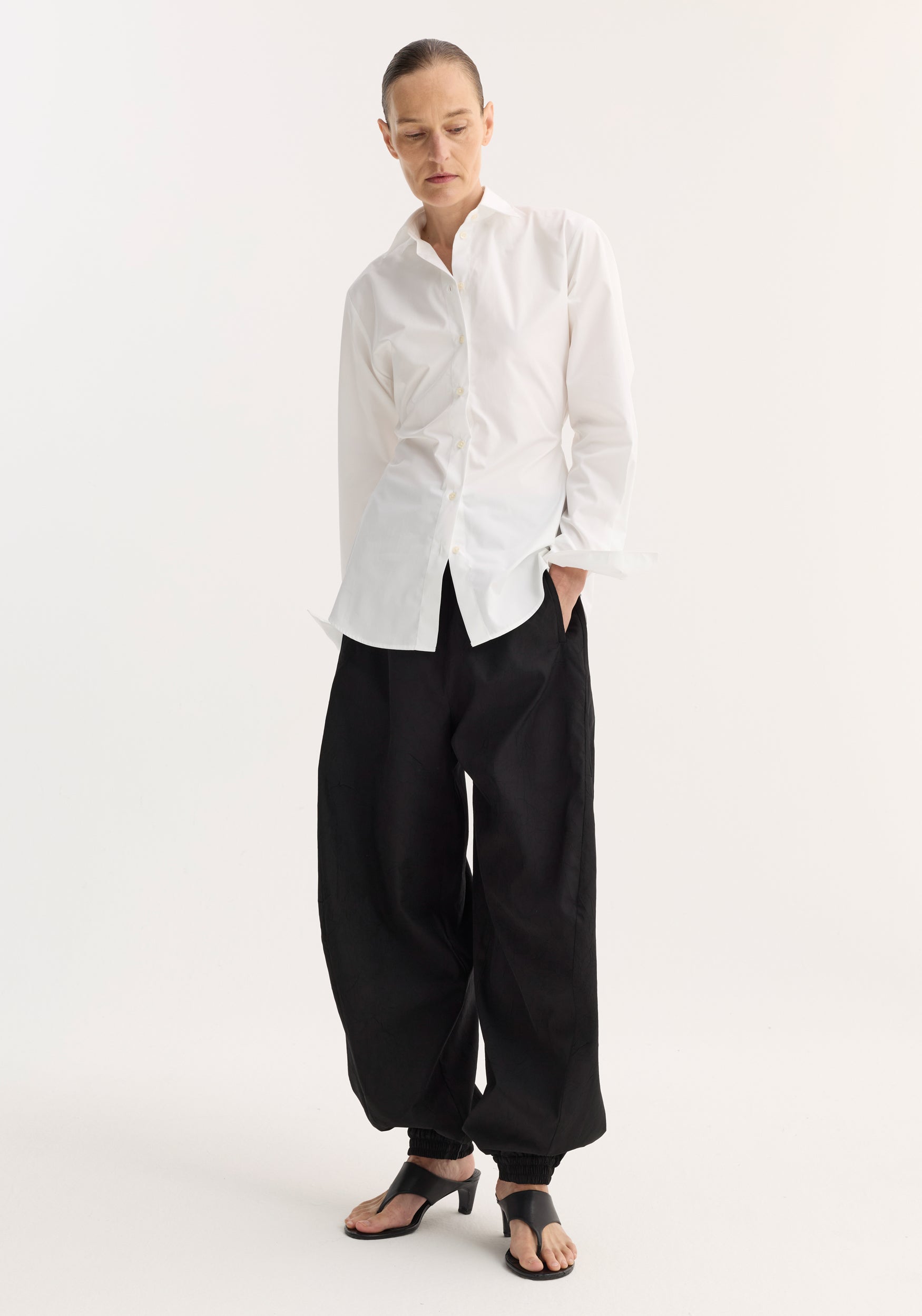 Person wearing a Poplin shaped shirt in optic white, featuring a relaxed hourglass fit and crafted from light cotton poplin, paired with loose black trousers.