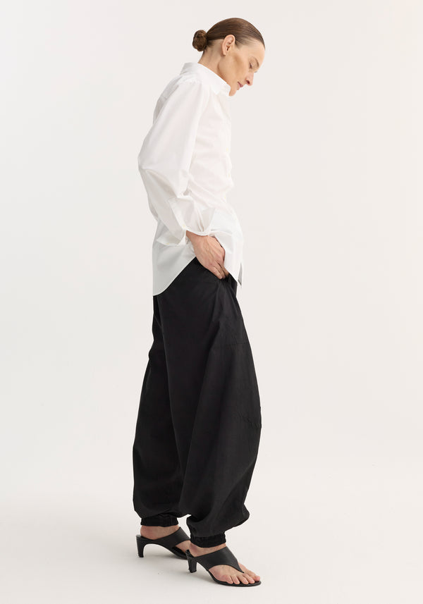 Model wearing Poplin shaped shirt | optic white, featuring a relaxed hourglass fit, untucked, paired with loose black pants and black slide sandals.