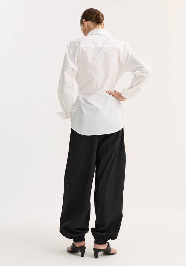 Model wearing a Poplin shaped shirt in optic white with a relaxed hourglass silhouette, paired with loose black pants and open-toe sandals.