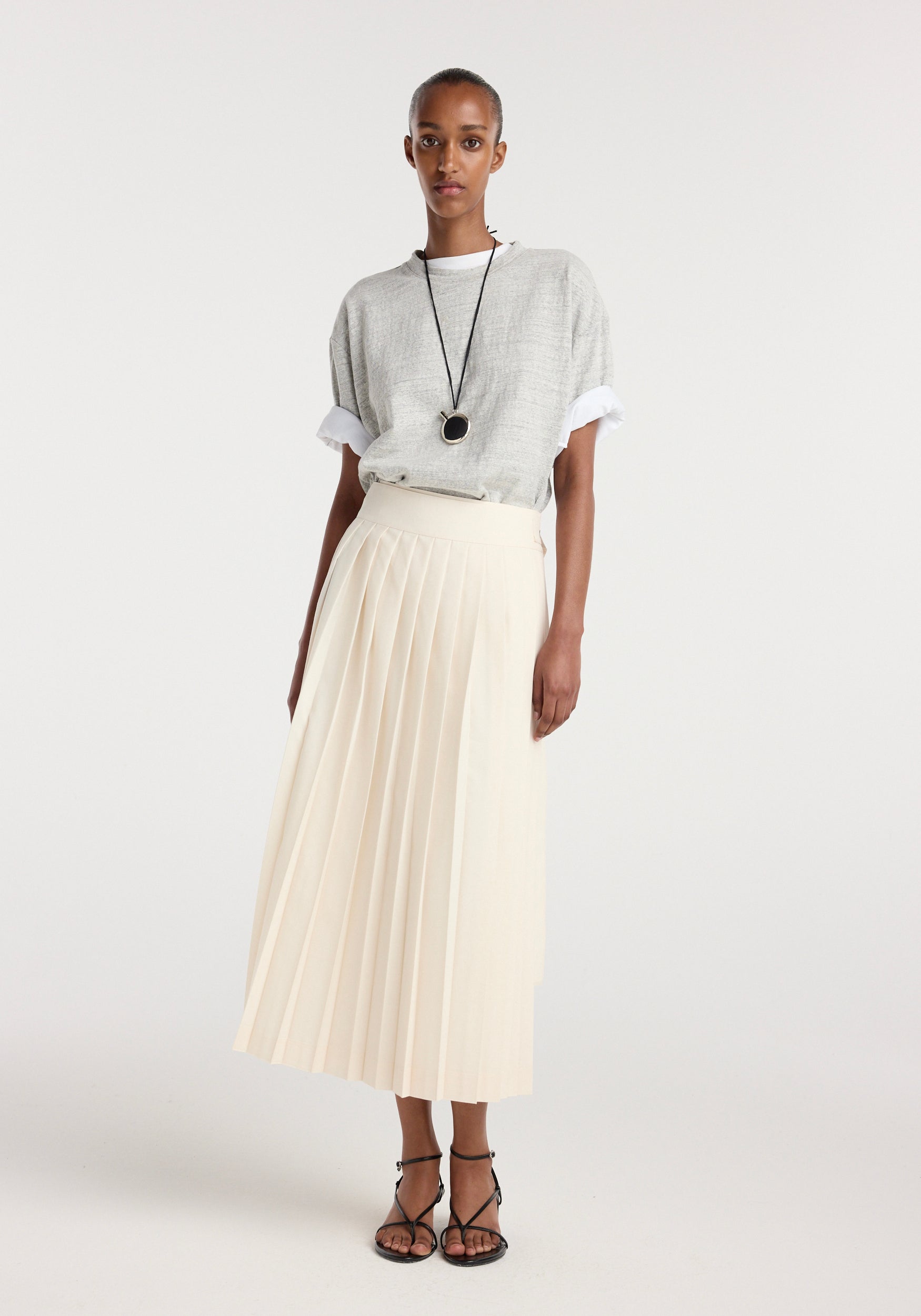 A woman wearing a cream plissé wrap skirt | cream paired with a casual grey top and black strappy sandals, accessorized with a long black pendant necklace.