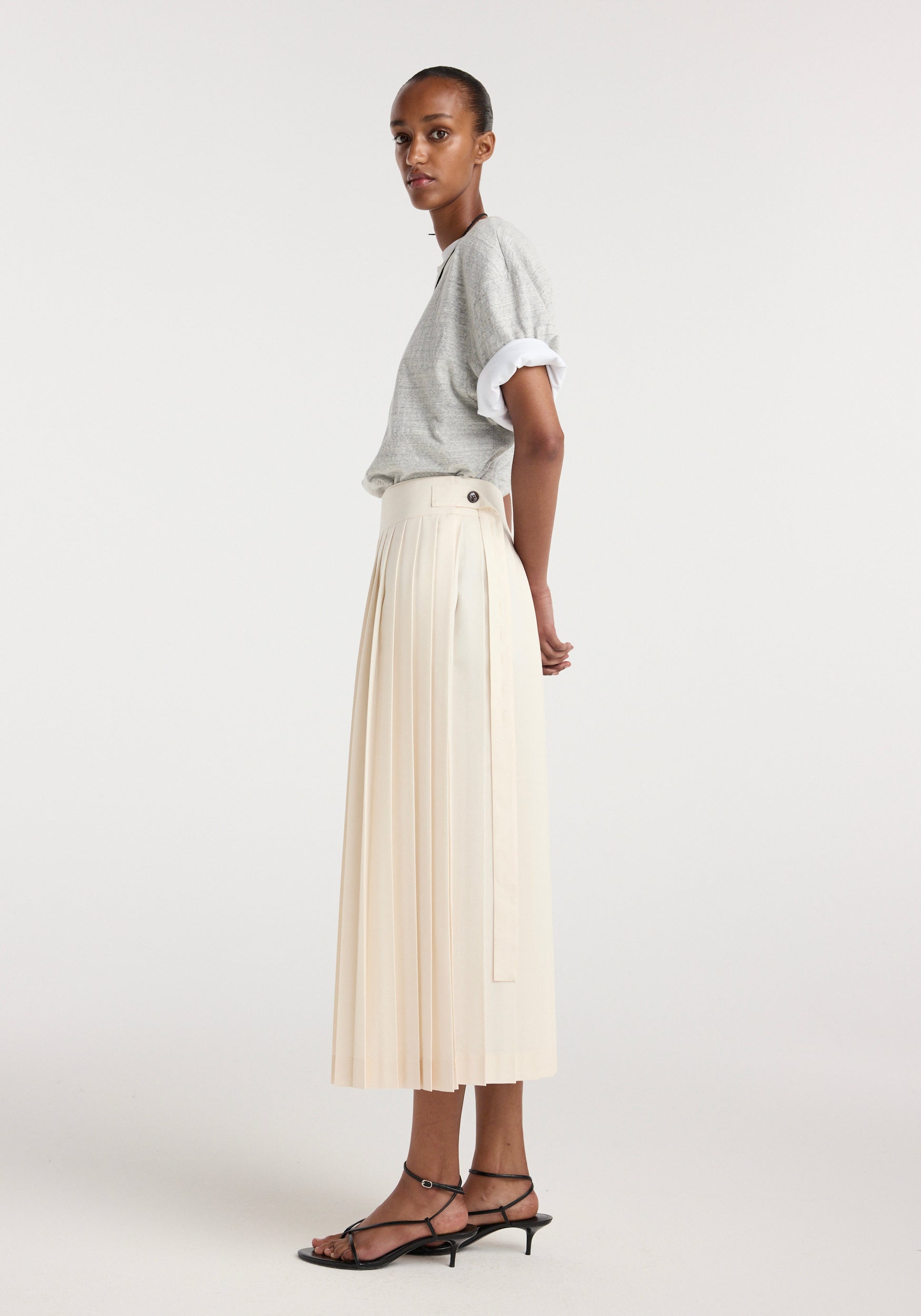 Model wearing a cream pleated midi-length Plissé wrap skirt with a side button detail, paired with a rolled-up short sleeve gray top and black strappy low-heeled sandals.