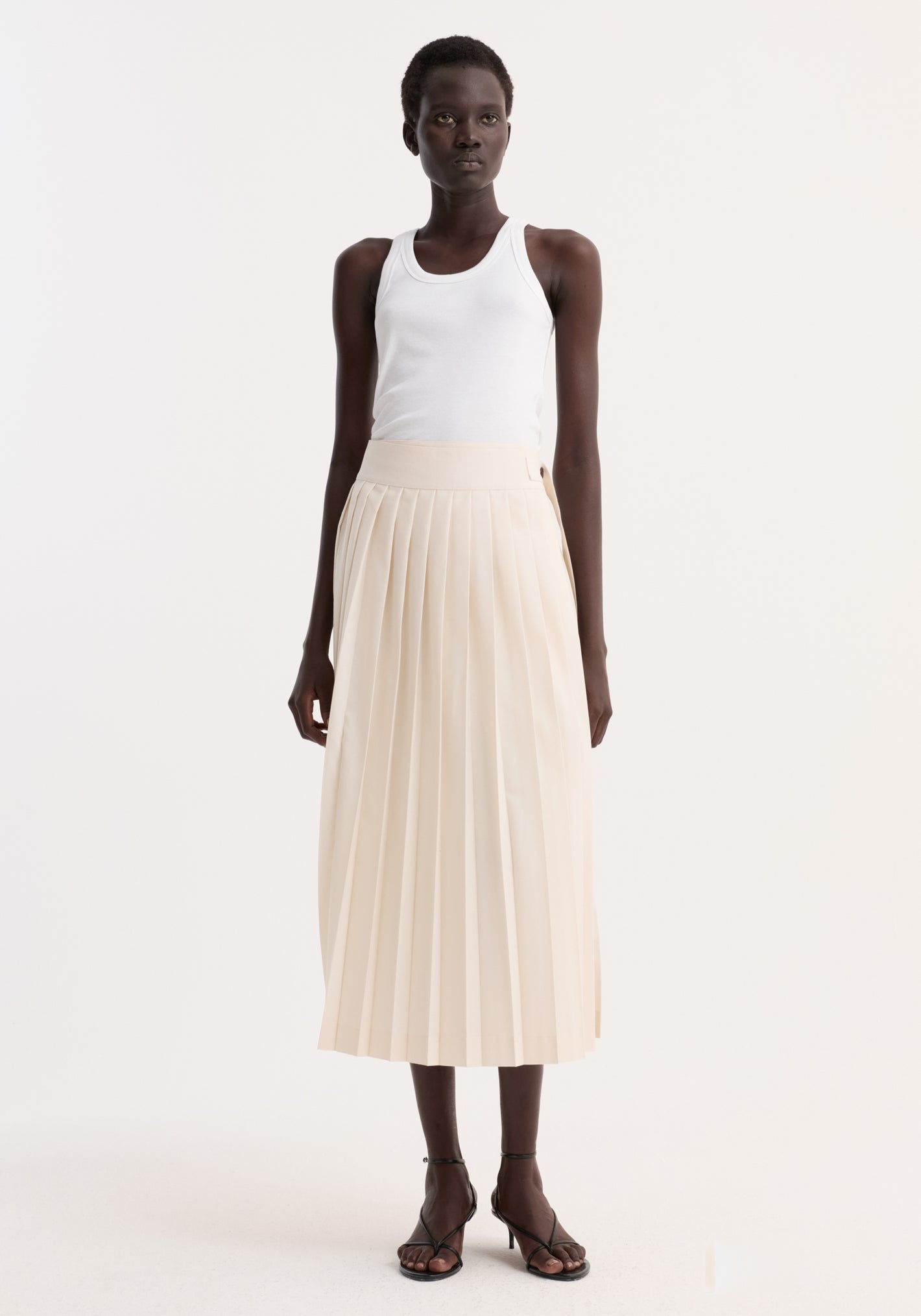 Model wearing Plissé wrap skirt | cream, featuring pleated wrap design, placket strap closure, and belt detail, styled with a white ribbed tank top and black sandals.