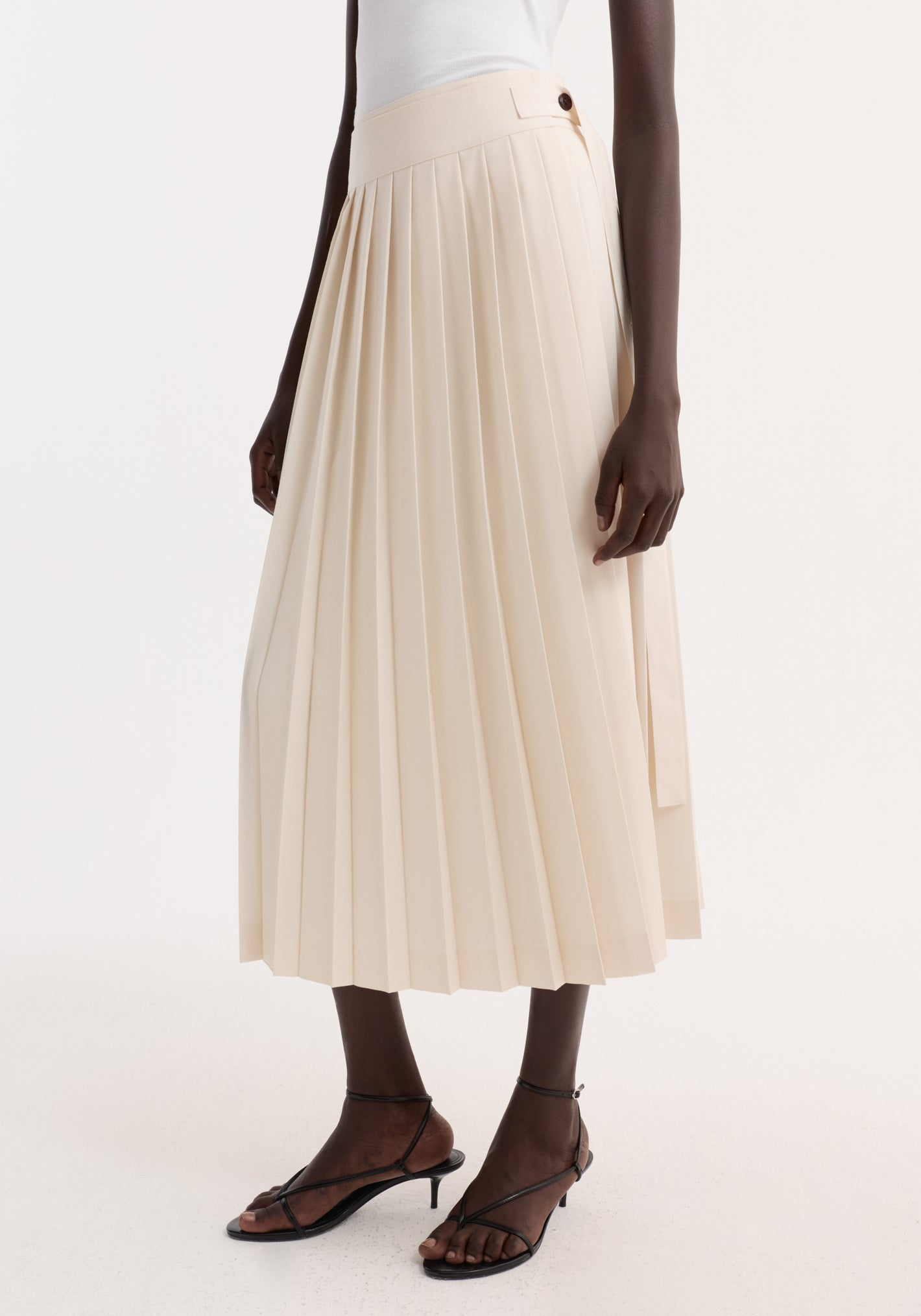 Model wearing a cream Plissé wrap skirt, featuring pleats, side-button detail, and a belt. Paired with black strappy heeled sandals.