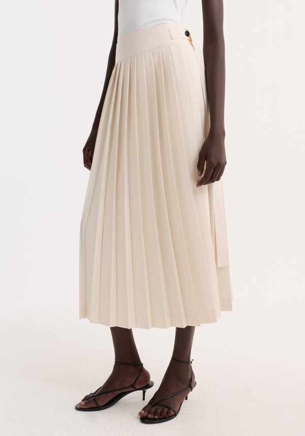 Model wearing a cream Plissé wrap skirt, featuring pleats, side-button detail, and a belt. Paired with black strappy heeled sandals.