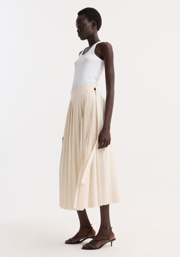 Woman models Plissé wrap skirt | cream, showcasing its pleated, wrap design with placket strap closure and belt detail, paired with a white tank top and black sandals.