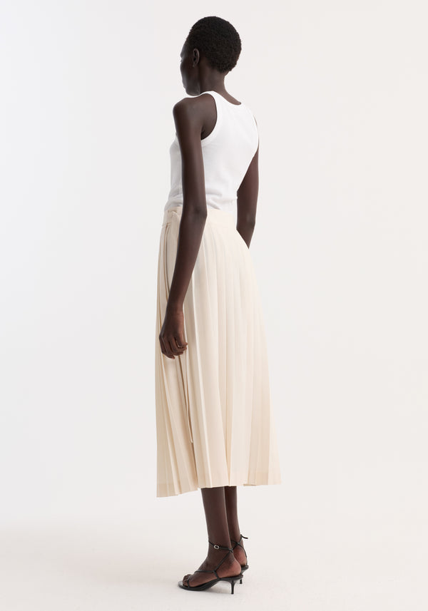 Model wearing Plissé wrap skirt in cream, featuring pleats and a wrap design with placket straps and belt, paired with a white tank top and black sandals.
