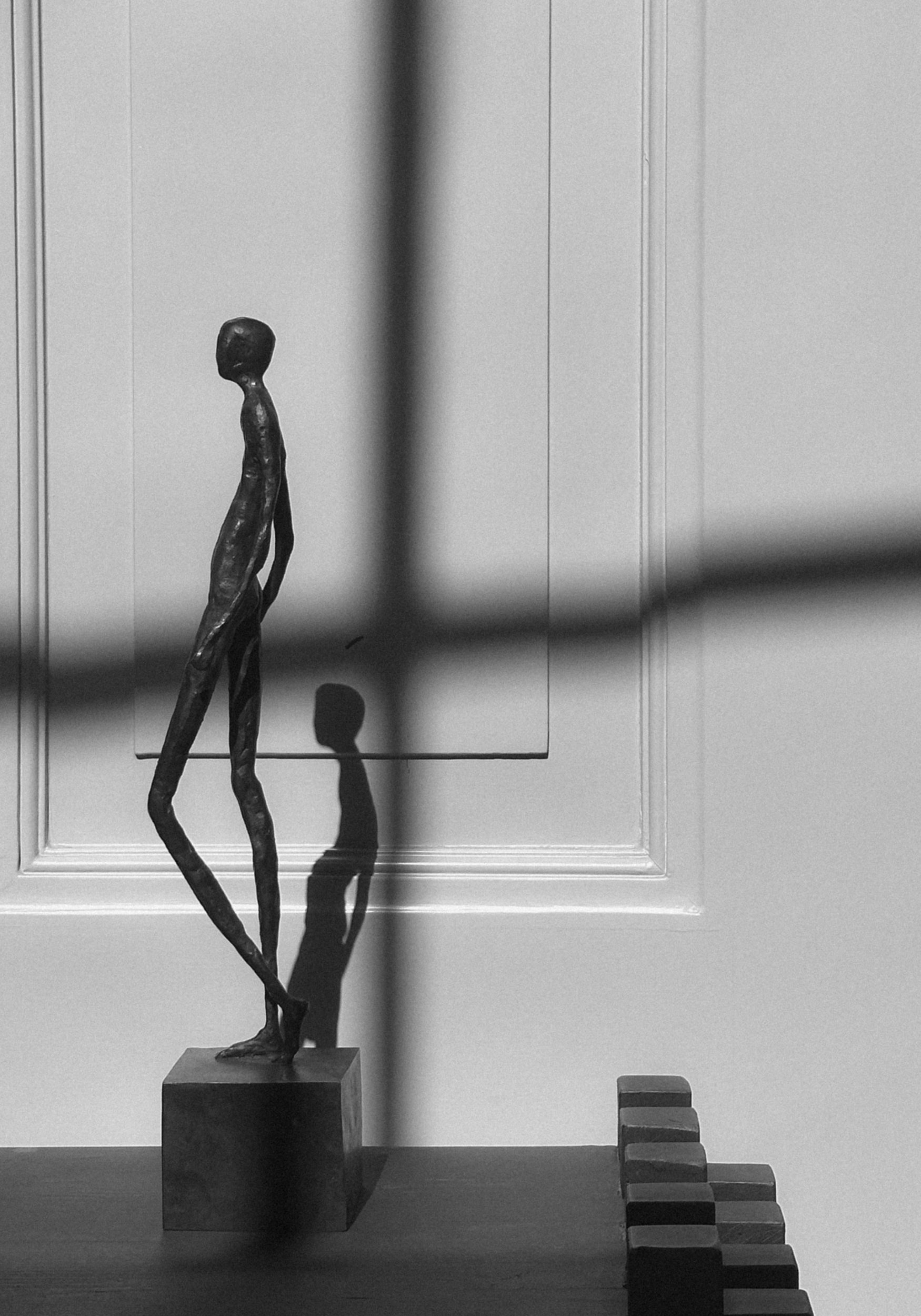Bronze Sculpture | Quiet Confidence: A slim, elongated bronze figure on a cuboid base, casting shadows on a paneled wall, handcrafted by Dutch artist Veri Wisman.
