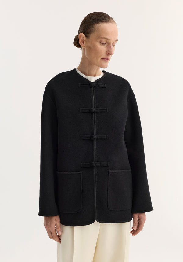 Model wearing the Mandarin double-faced wool jacket in black, featuring a mandarin collar, frog toggles, embroidered edges, and hip-level patch pockets.