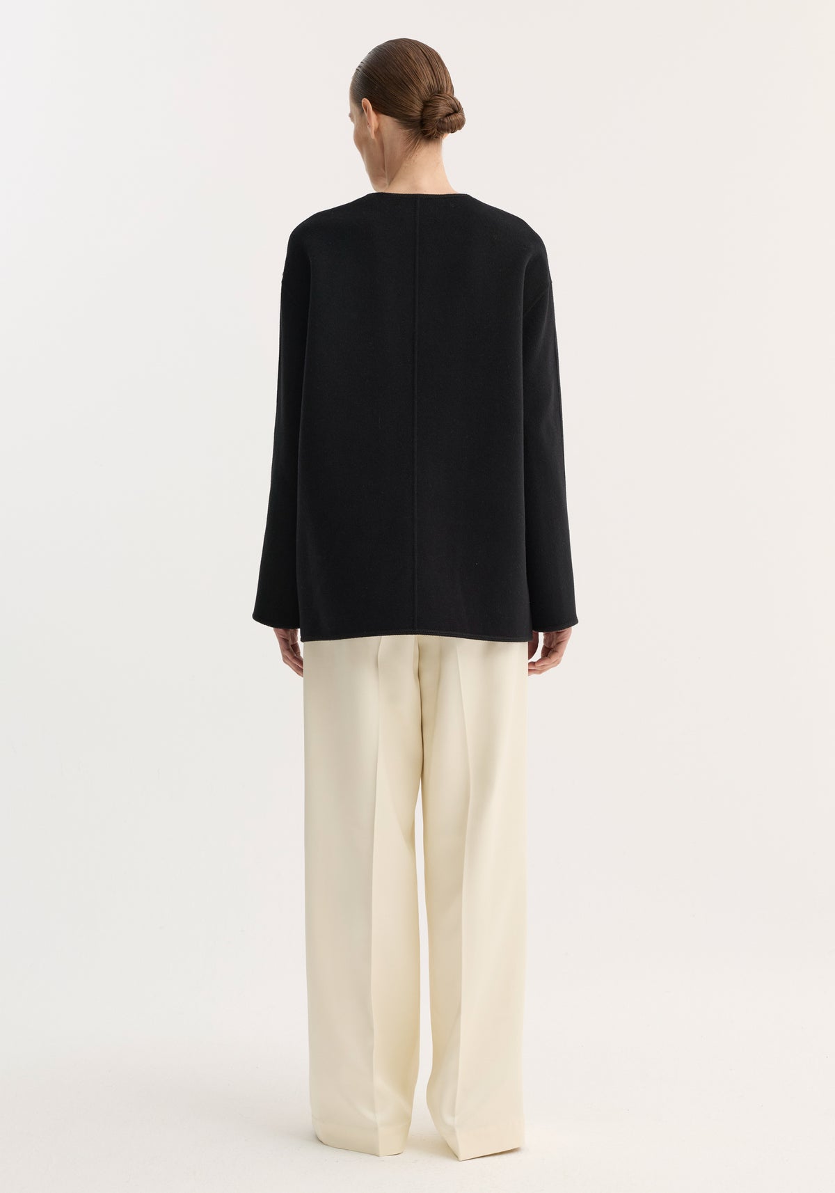 Rear view of person wearing a loose-fitting black top over cream trousers, showcasing the Mandarin double-faced wool jacket with embroidered edges and patch pockets.
