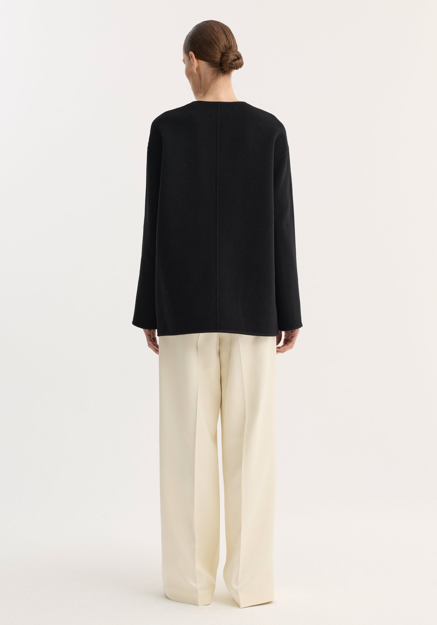 Rear view of person wearing a loose-fitting black top over cream trousers, showcasing the Mandarin double-faced wool jacket with embroidered edges and patch pockets.