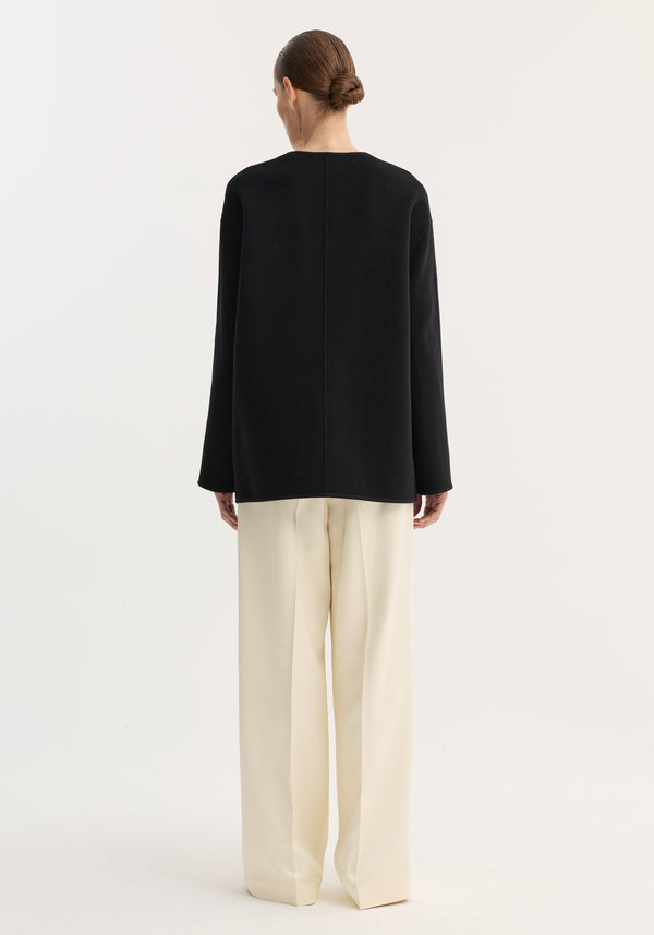 Rear view of person wearing a loose-fitting black top over cream trousers, showcasing the Mandarin double-faced wool jacket with embroidered edges and patch pockets.