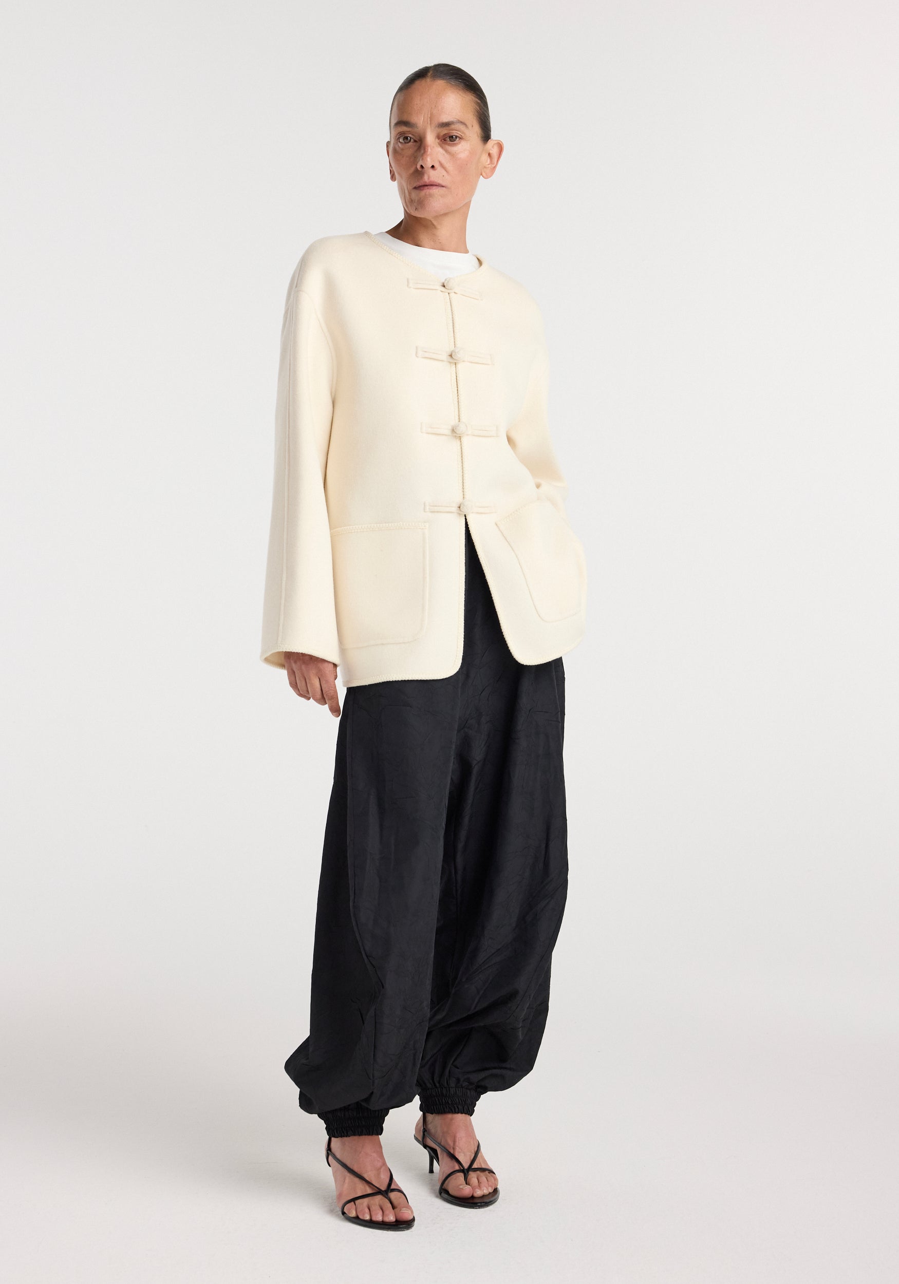 Woman wearing a double-faced wool pankou jacket in off white with traditional toggle fastenings, paired with loose black pants and black strappy heels, standing against a plain background.