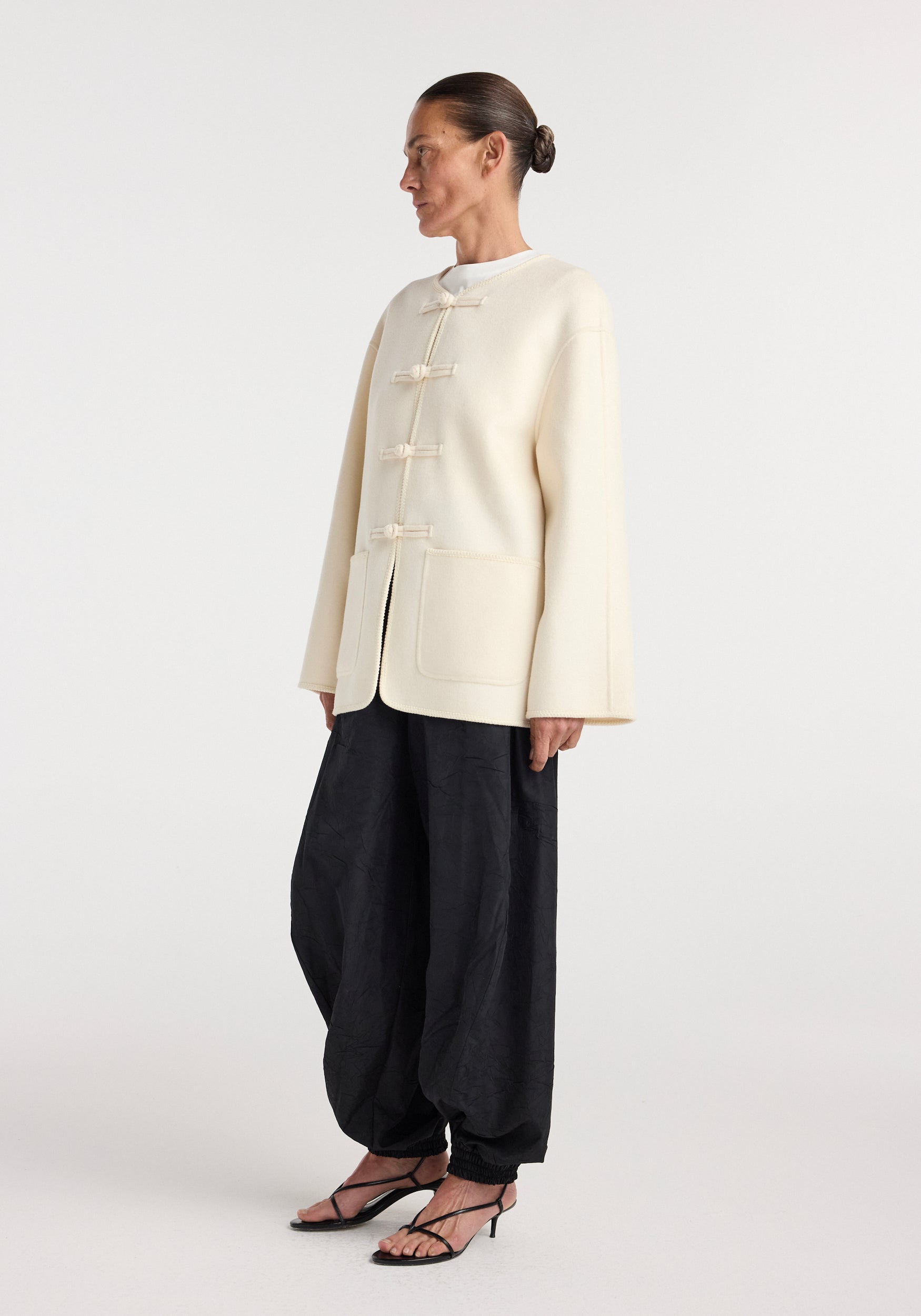 Side view of a model wearing a Double-faced wool pankou jacket | off white with traditional pankou knot buttons, paired with loose black pants and strappy heels.