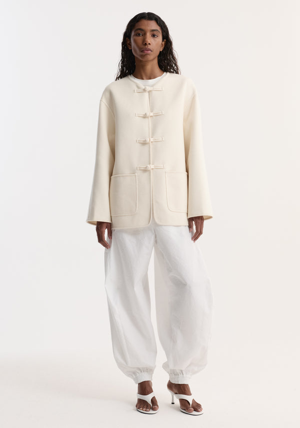 Model wears Double-faced wool pankou jacket with embroidered edges and patch pockets, fastened with traditional pankou knots, paired with loose, cuffed trousers and heeled sandals.