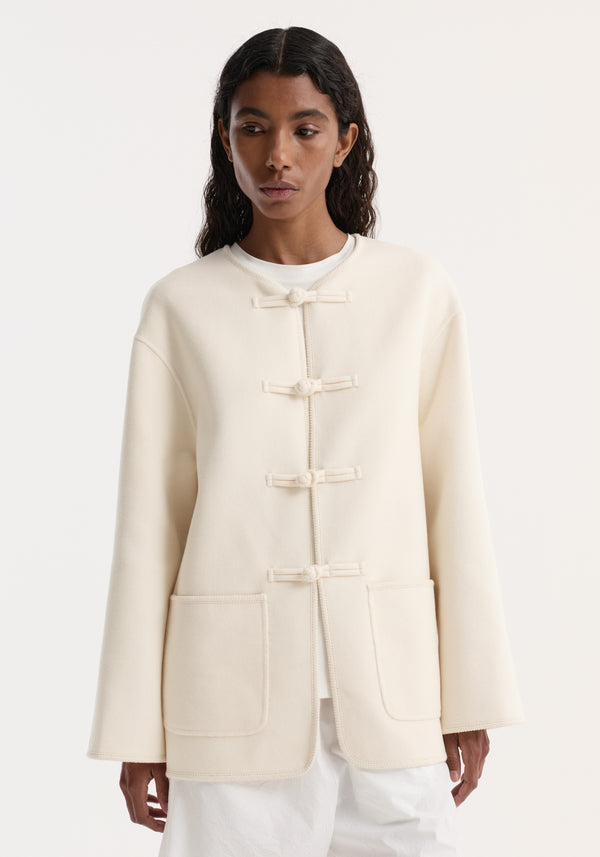 Woman wearing double-faced wool pankou jacket in off white, featuring embroidered edges, patch pockets, and traditional knotted closures, creating a minimalist silhouette.