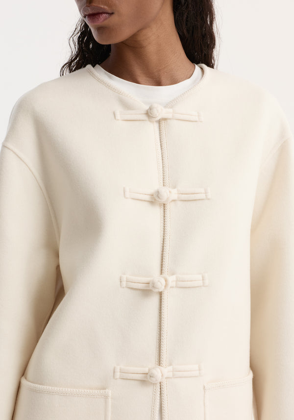 Close-up of a woman wearing a double-faced wool pankou jacket with frog closures, embroidered edges, and patch pockets, layered over a plain white T-shirt.