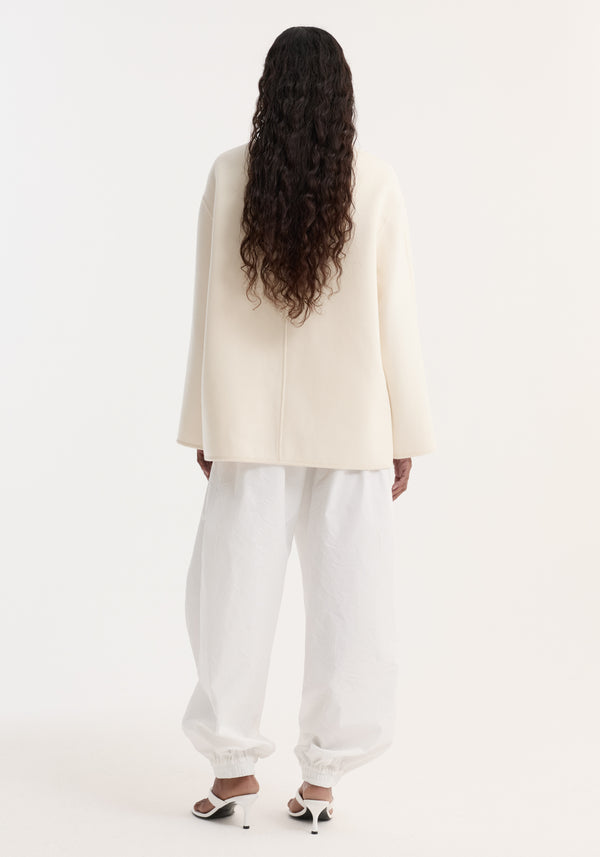 Rear view of a woman in a Double-faced wool pankou jacket, featuring embroidered edges and patch pockets, paired with white wide-leg trousers and strappy heeled sandals.