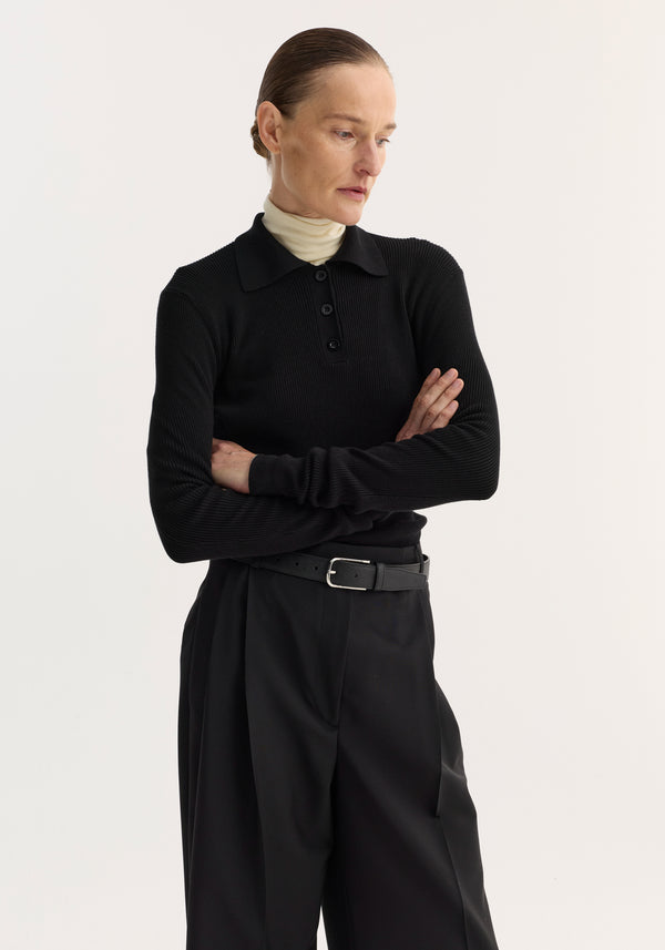 Person wearing a Rib knitted polo | black over a cream turtleneck, styled with high-waisted trousers and a slicked-back hairstyle, exuding a minimalist, editorial look.