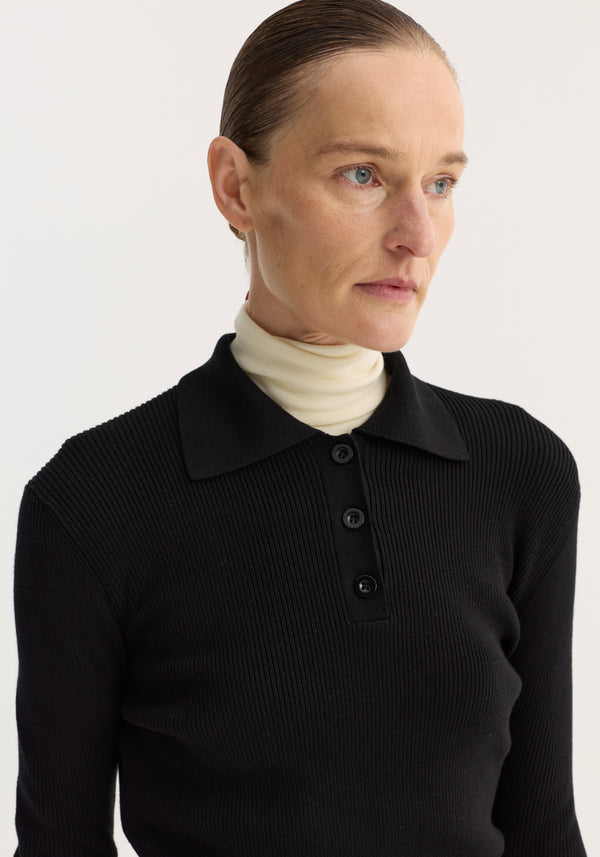 Person wearing Rib knitted polo | black, featuring a ribbed texture, three-button neckline, and long sleeves, layered over a light turtleneck.