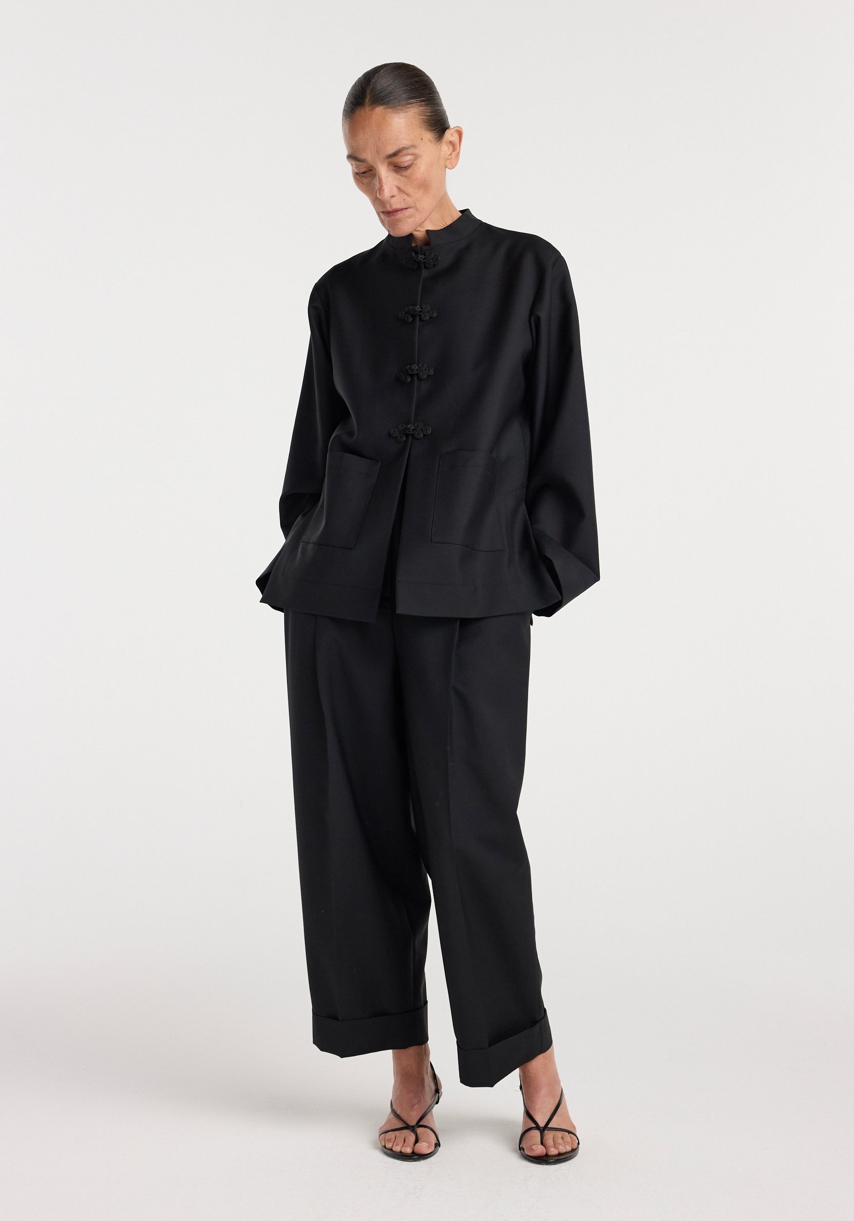 Woman wearing a black Pankou closure jacket | black with traditional frog buttons, paired with loose black trousers and black strappy heels against a plain white background.