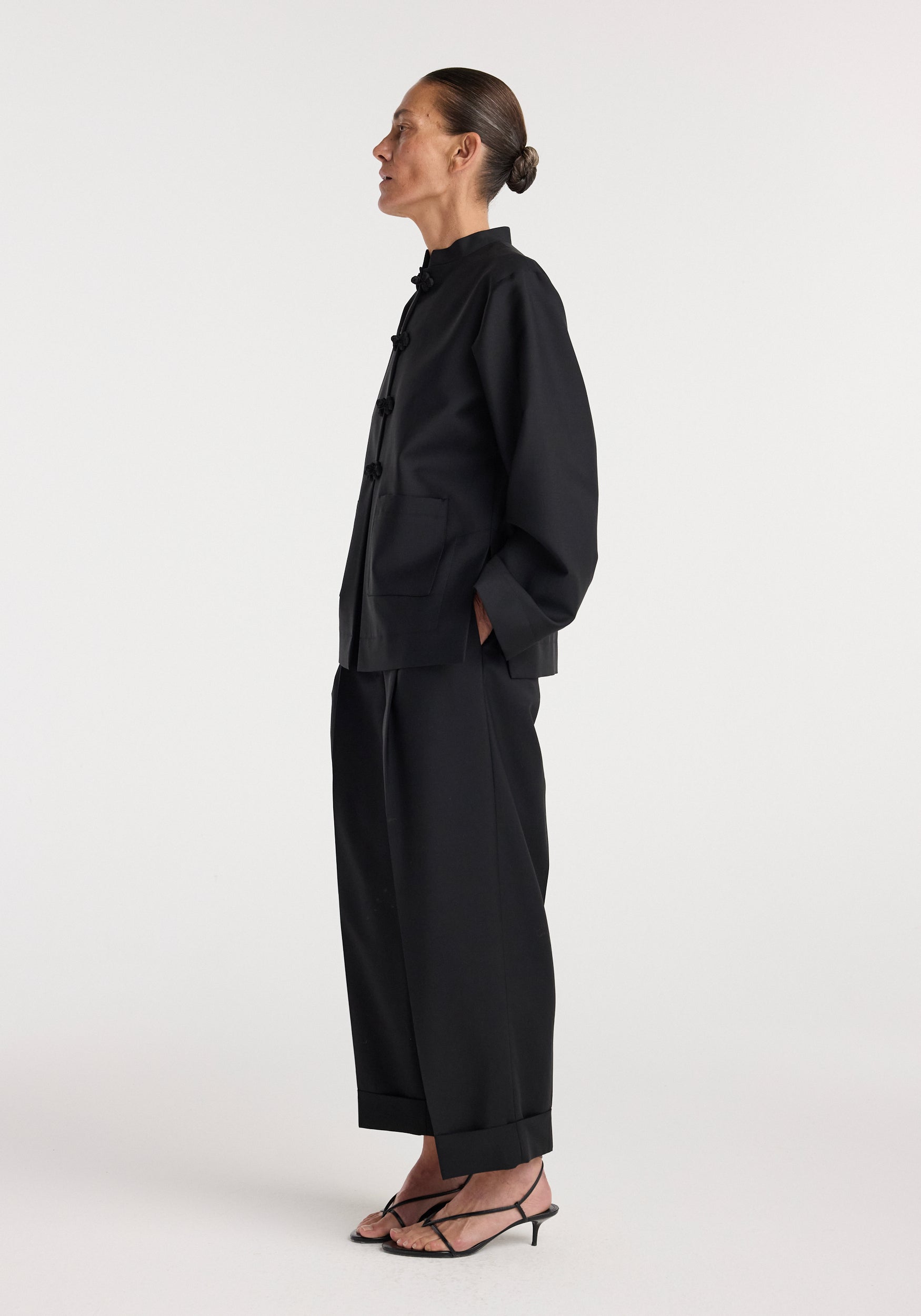 Side view of a person wearing a black Pankou closure jacket | black with a mandarin collar, loose fit sleeves, and front pockets paired with black wide-leg pants and black open-toe heels.