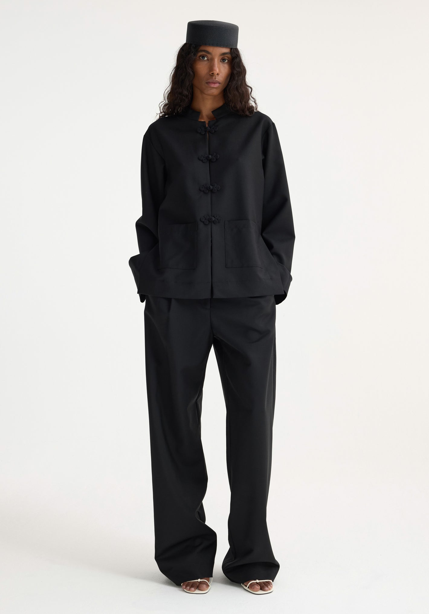 Model wears Mandarin jacket | black with frog closures, loose trousers, and pillbox hat, showcasing a minimalist take on traditional Chinese tailoring.