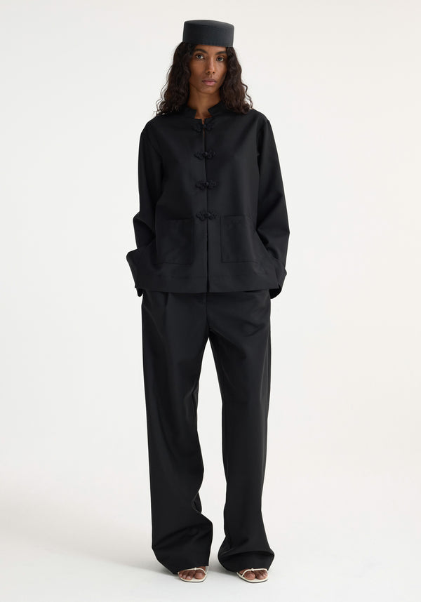 Model wears Mandarin jacket | black with frog closures, loose trousers, and pillbox hat, showcasing a minimalist take on traditional Chinese tailoring.