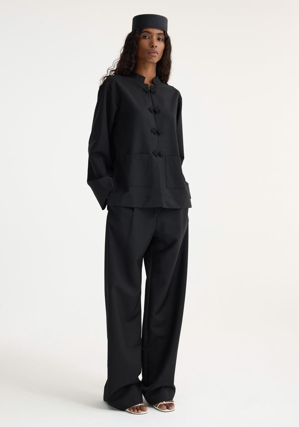 Woman wearing the Mandarin jacket | black with frog-style closures, patch pockets, paired with wide-leg trousers and a pillbox cap for a minimalist look.