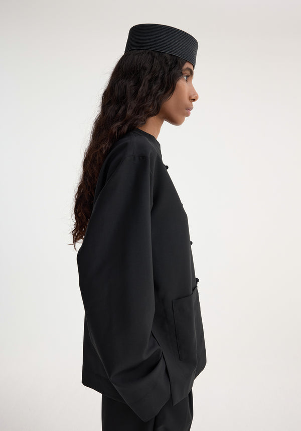 Person in side profile wearing a black Mandarin jacket with patch pockets and pankou closure, complemented by a matching hat and trousers.