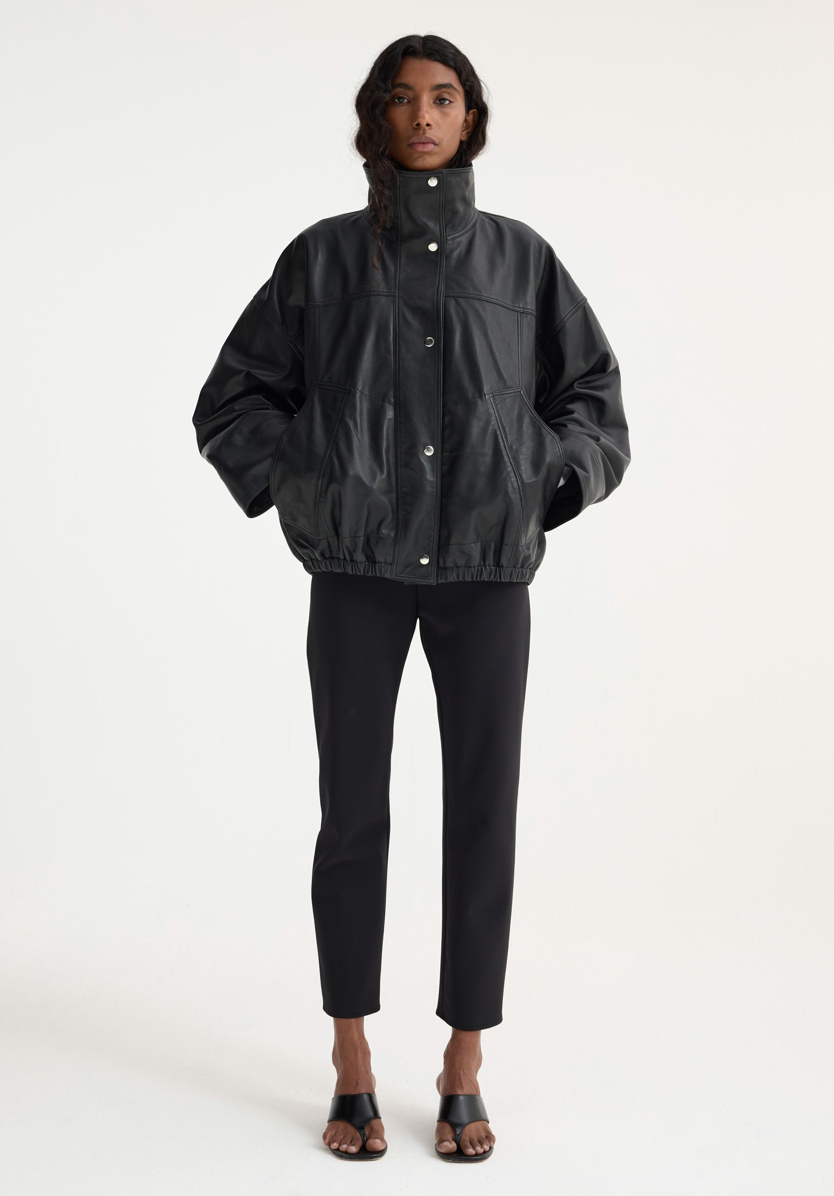 Model in boxy Reinvented leather jacket | black, with high collar and snap closures, paired with fitted trousers and mule sandals against a white backdrop.