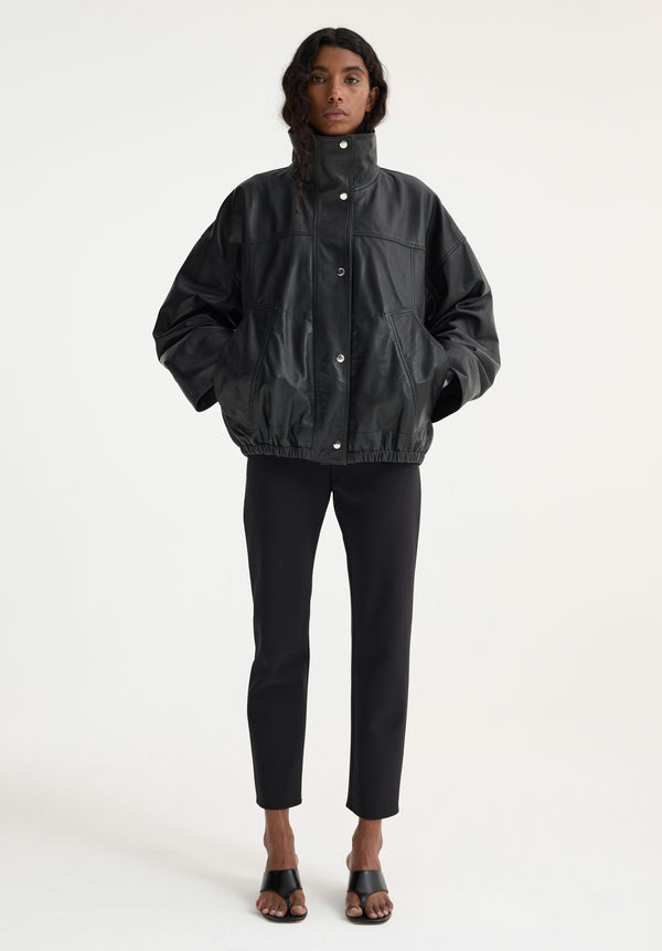 Model in boxy Reinvented leather jacket | black, with high collar and snap closures, paired with fitted trousers and mule sandals against a white backdrop.