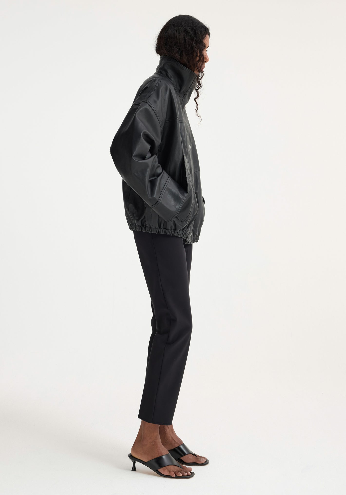 Model wears an oversized, soft leather, black Reinvented leather jacket with a high collar and elastic hem, hands in pockets, styled with black trousers and sandals.