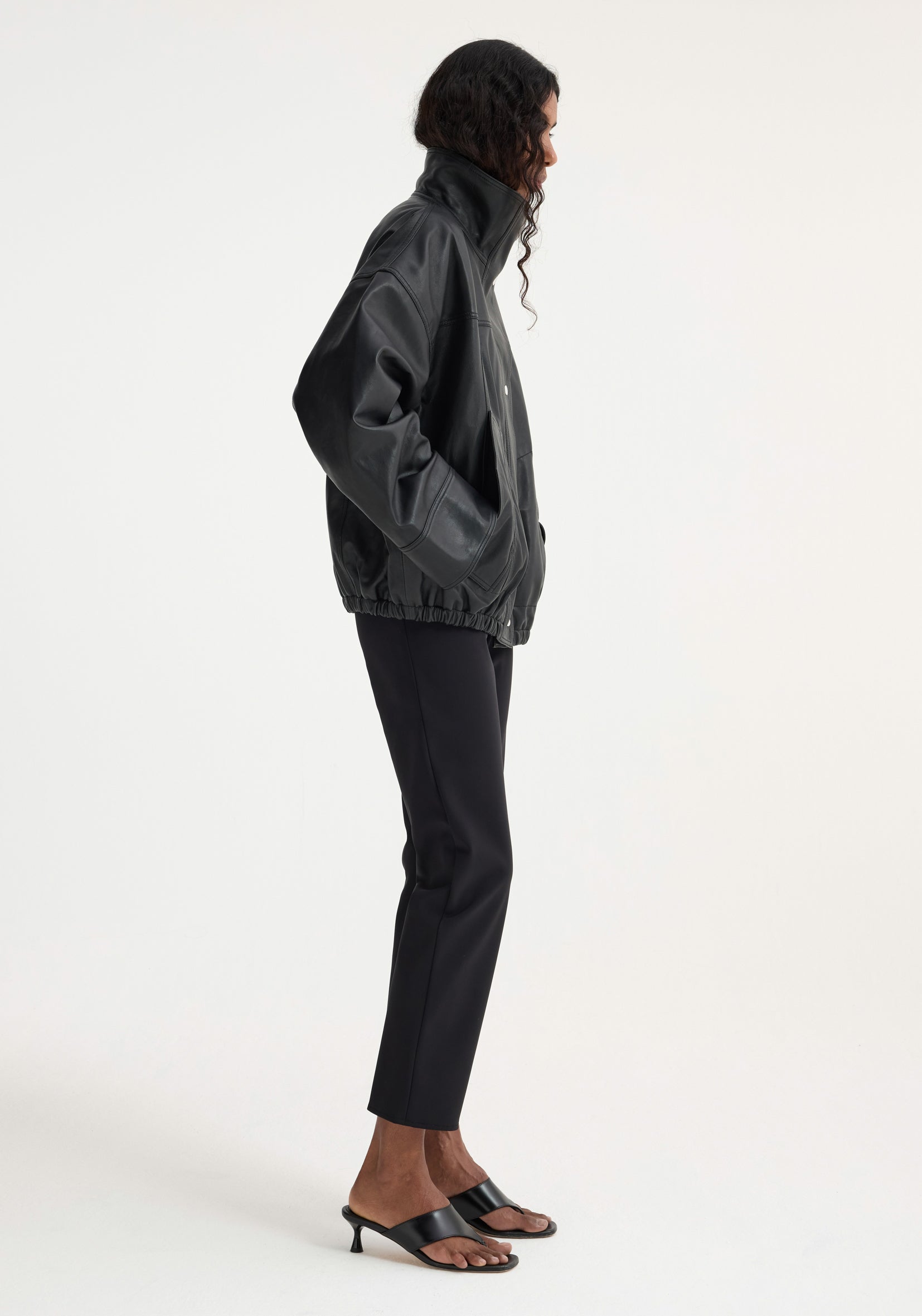 Model wears an oversized, soft leather, black Reinvented leather jacket with a high collar and elastic hem, hands in pockets, styled with black trousers and sandals.