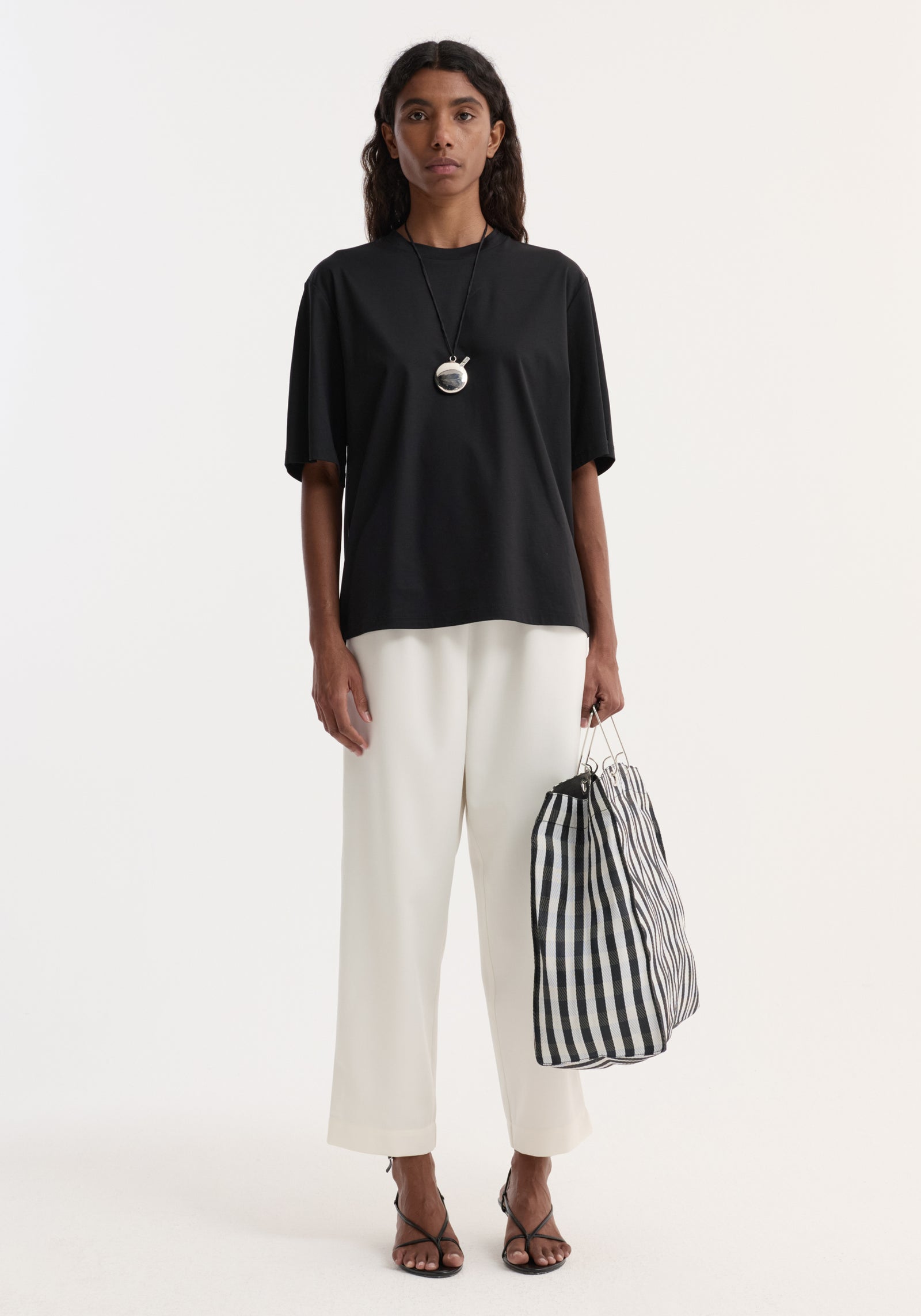 Person modeling the Oversized cotton t-shirt | black, featuring an oversized fit and drop shoulder, paired with wide-leg trousers and a checked tote bag.