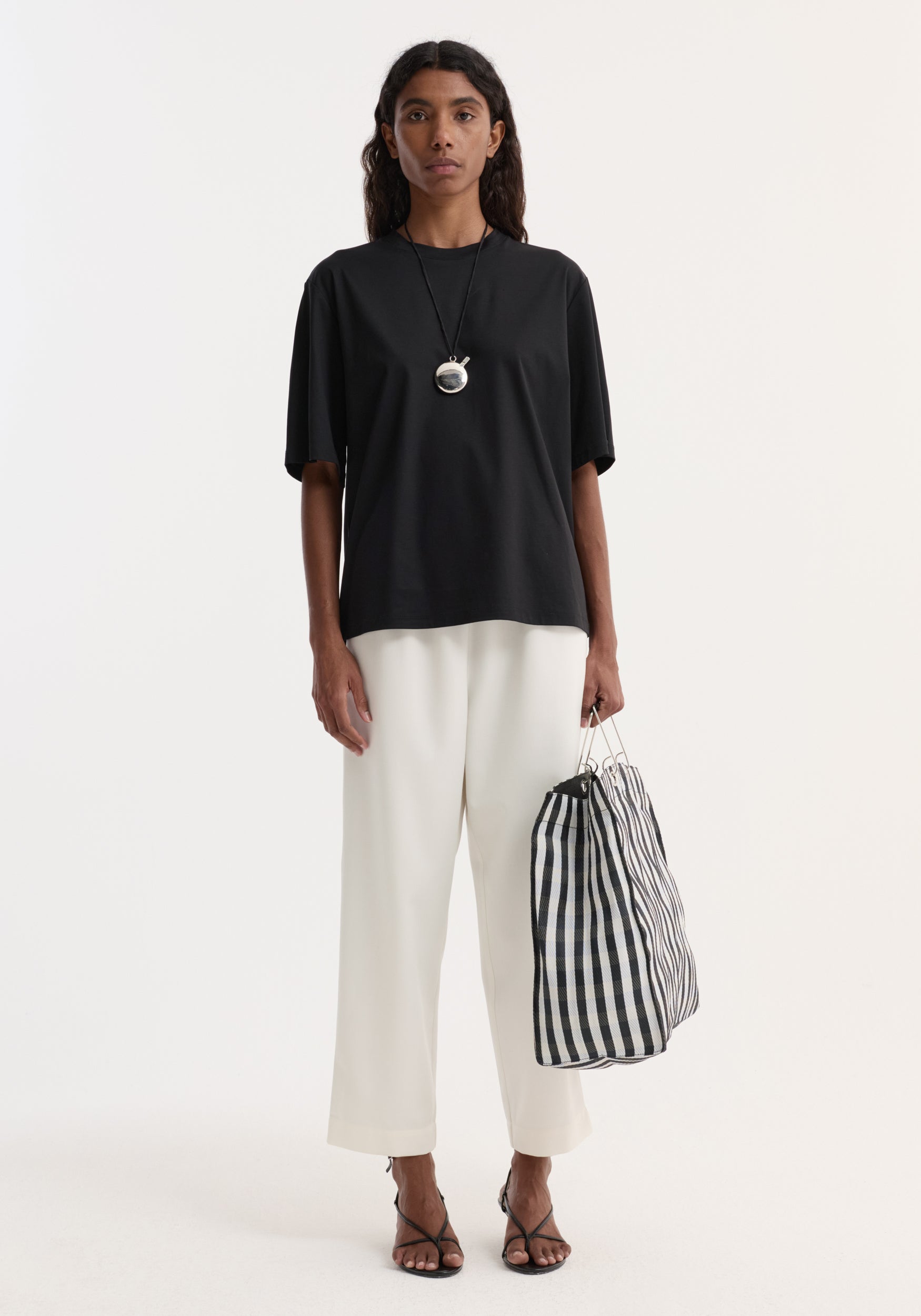Woman wearing a Silver Perfume Bottle Necklace, a black T-shirt, white trousers, black sandals, and holding a gingham tote, showcasing minimalist style.