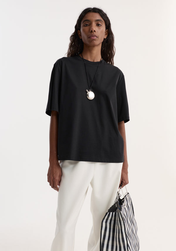 Model wearing the Oversized cotton t-shirt | black, featuring a loose fit with a crew neck, styled with white trousers and a checkered tote bag.