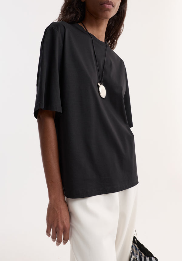 Person wearing an Oversized cotton t-shirt in black, showcasing its relaxed fit, crew neck, and dropped shoulders, paired with a silver pendant necklace.
