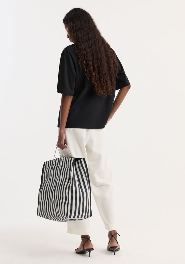 Rear view of a person in an Oversized cotton t-shirt | black, tucked into white trousers, holding a striped tote, highlighting the t-shirt's relaxed fit and crew neck.
