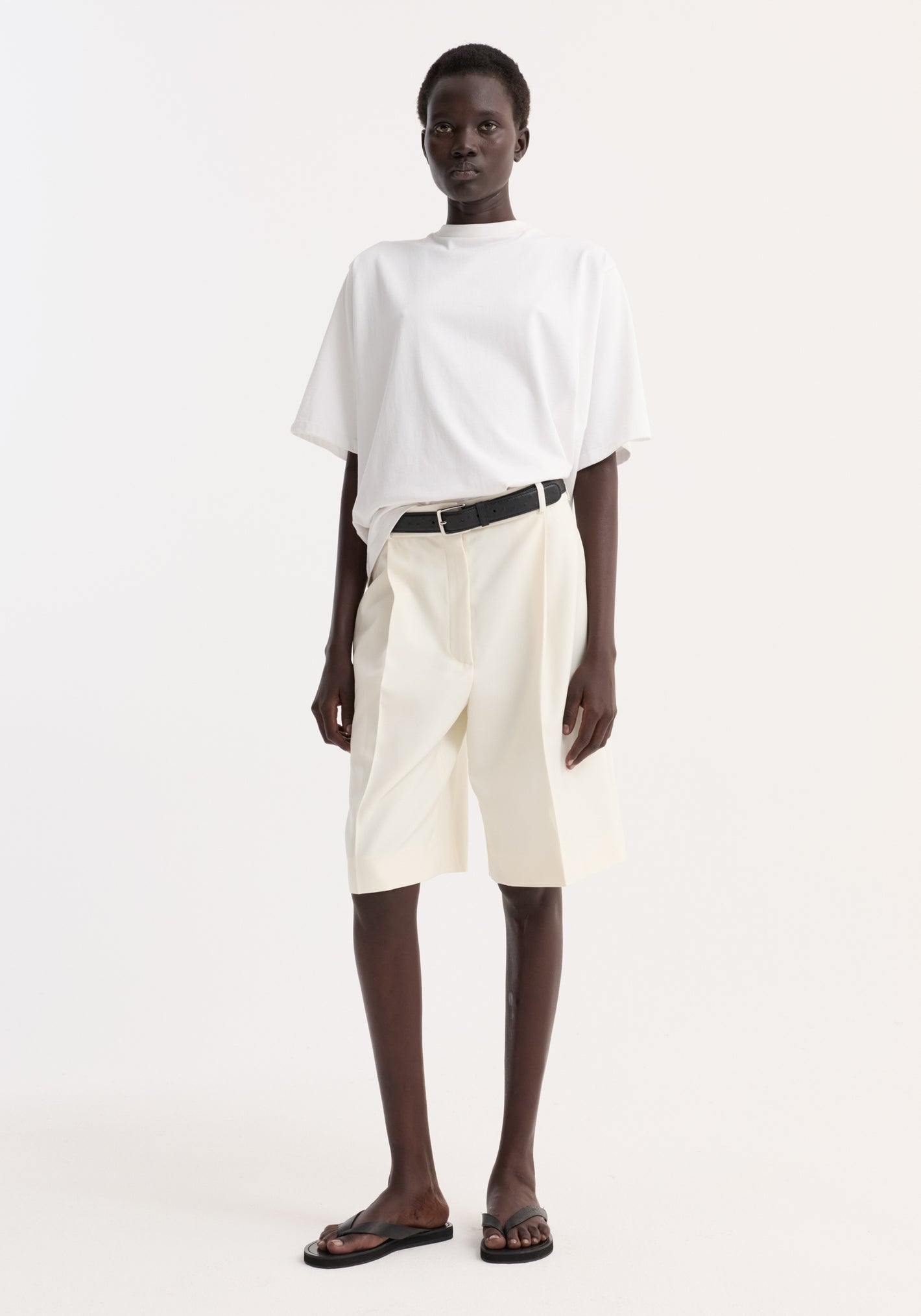 Model wearing an Oversized cotton t-shirt | optic white with high-waisted shorts, black leather belt, and black thong sandals, showcasing a relaxed, polished style.