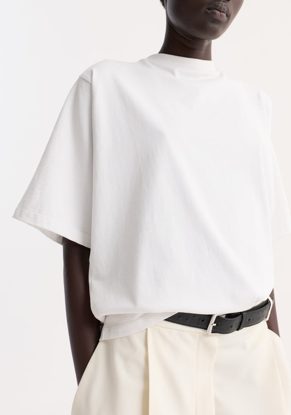 Person wearing an Oversized cotton t-shirt | optic white, tucked into high-waisted trousers, featuring a relaxed fit, drop shoulder, crew neck, and brand logo on sleeve.
