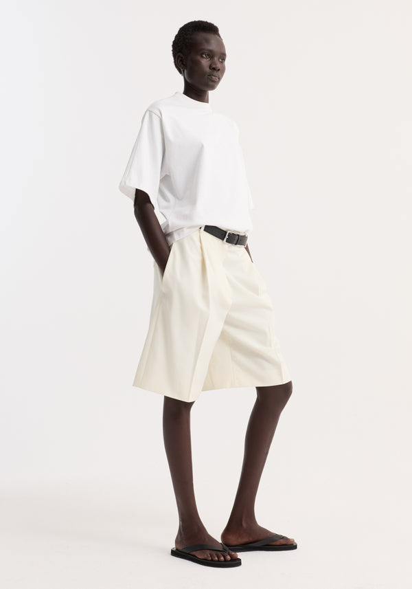 Model wearing Oversized cotton t-shirt in optic white; features relaxed fit, drop shoulder, crew neck, and side seam logo, tucked into pleated shorts.