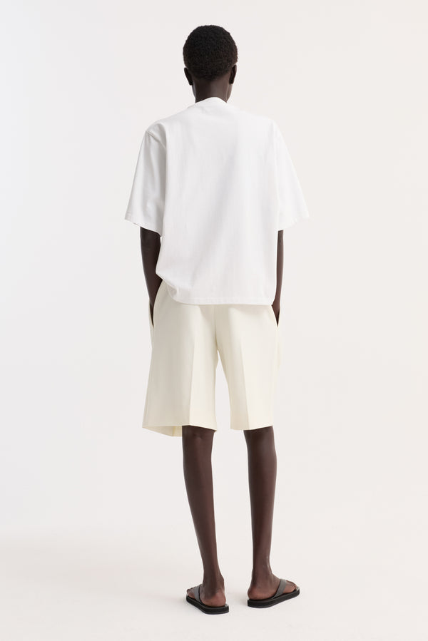 Back view of person wearing Oversized cotton t-shirt in optic white with dropped shoulder, paired with tailored shorts and black sandals, hands in pockets.